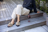 Woman wearing Baza Shearling slide sandal in bronze/black with beige pants and sweater, next to wine glass