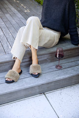 Woman wearing Baza Shearling slide sandal in bronze/black with beige pants and sweater, next to wine glass