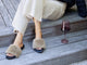 Woman wearing Baza Shearling slide sandal in bronze/black with beige pants and sweater, next to wine glass