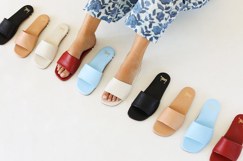 Woman wearing Honeybird leather slide sandals in red and eggshell with floral pants lined up with Honeybird sandals in black and beach.