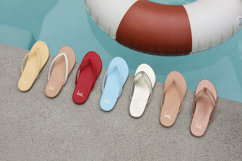 Group shot of Sunbeam leather flip flop sandals in glow, vanilla/beach, lipstick, sky, platinum, tan, and rose gold by the pool